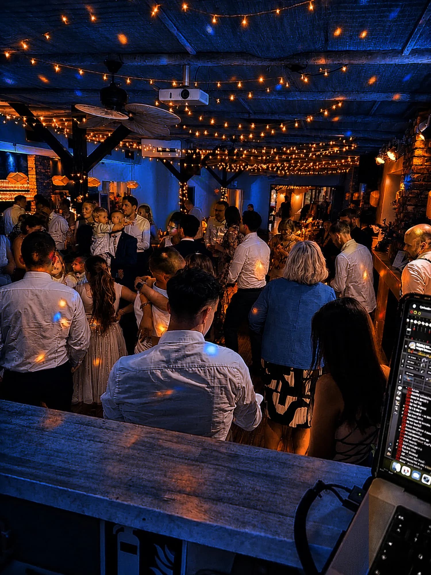 Blick vom DJ-Pult über einen vollen Raum mit Lichterketten, Gäste im warmen und blauen Licht